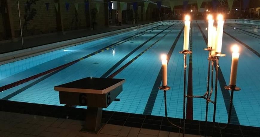 En ljusstake med levande ljust står i förgrunden. I bakgrunden en stämningsfullt upplyst simbassäng och lokalen är nedsläckt.
