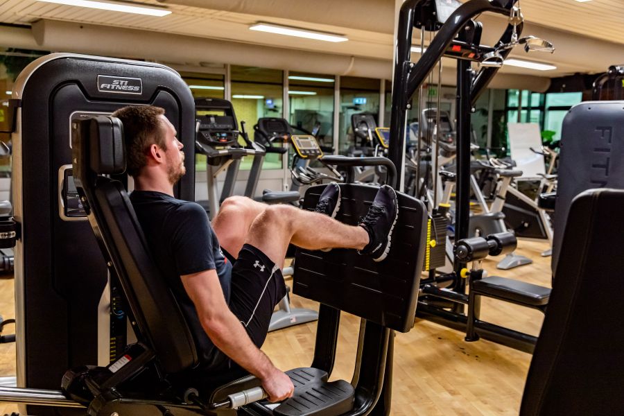 En man som tränar benpress i en maskin på ett gym. I bakgrunden syns flera träningsmaskiner.