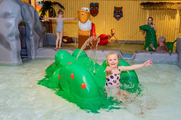 En liten flicka i prickig baddräkt åker rutschkana ner för en stor grön groda som sprutar vatten. I bakgrunden syns flera leksaker. 
