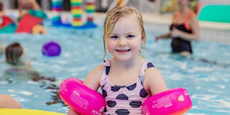 En liten flicka med rosa armpuffar och prickig baddräkt badar i en bassäng